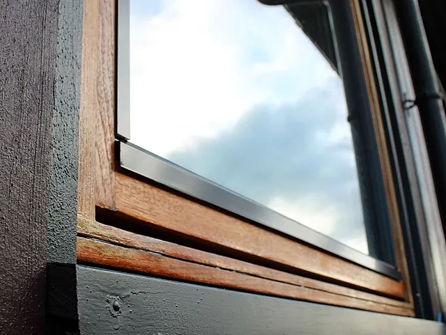 Nærbilde av vinduskarm i tre og metall, som viser detaljert finish og speiling i glasset.